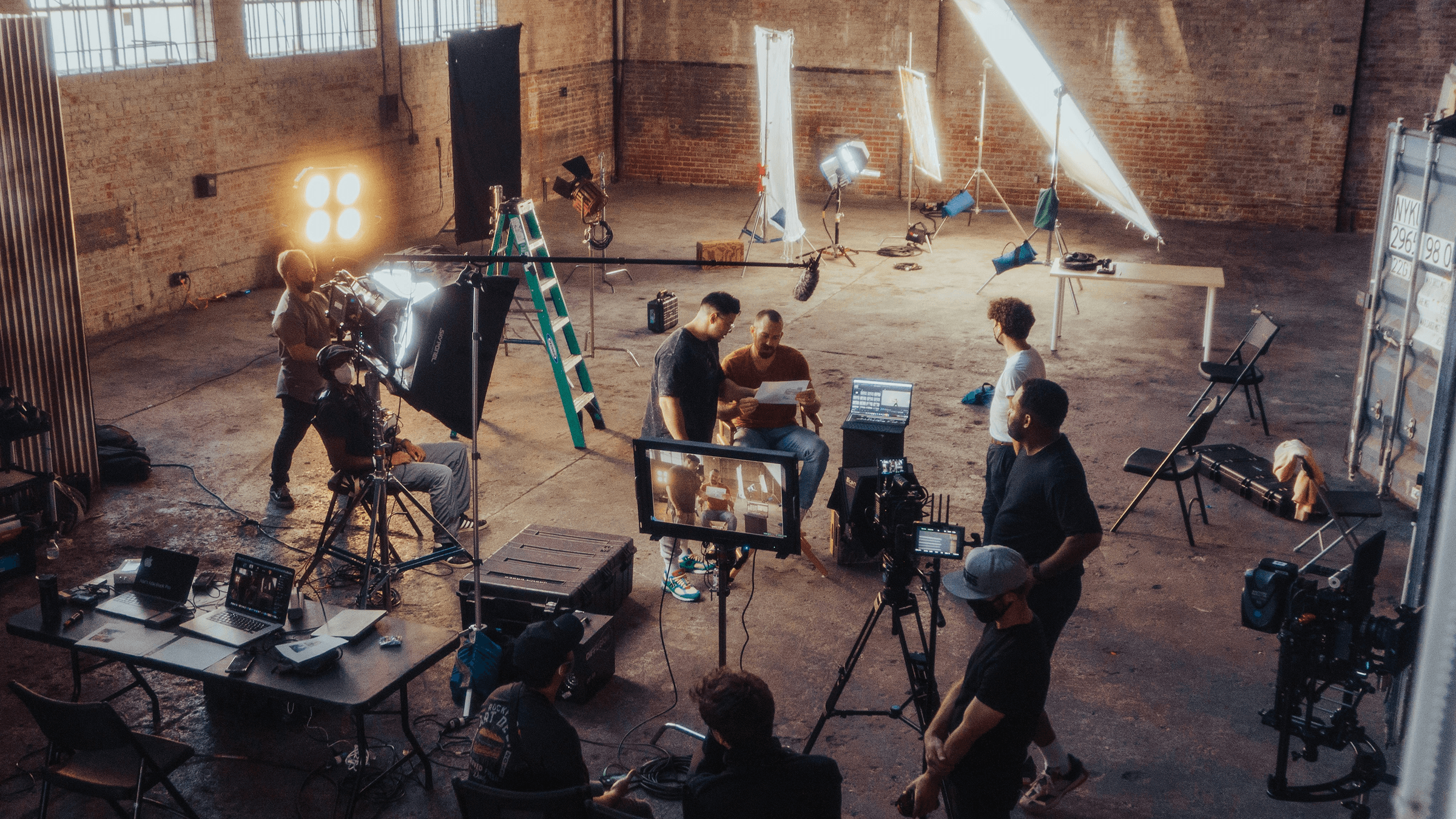 Film crew working on a lit brick-warehouse set, with cameras, lights and a monitor showing the shot, a picture of the work behind an experience that the audience never sees